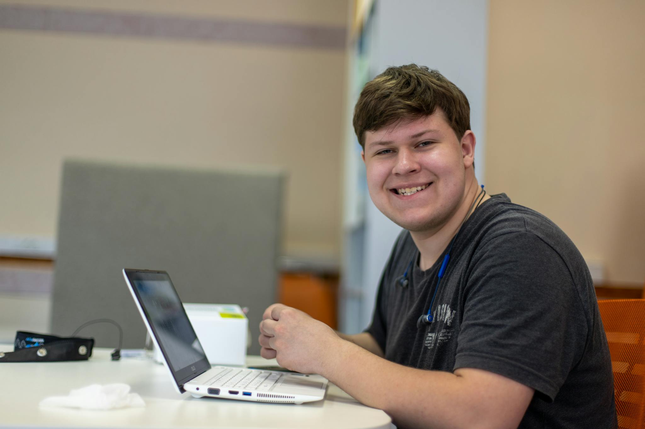 Young man smiling while working on a laptop in a modern office setting, showing relaxed and productive atmosphere.